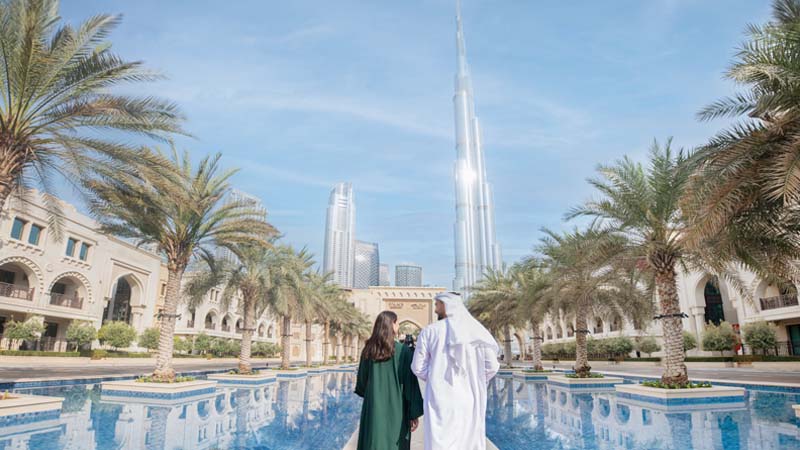 People walking along the pool and palm trees, with tall buildings in the background