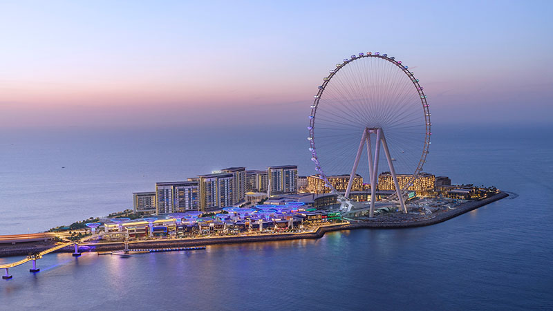 Aerial view of a coastal cityscape at dusk, featuring a large Ferris wheel and modern buildings on an island, with calm blue sea and pink-tinged sky.
