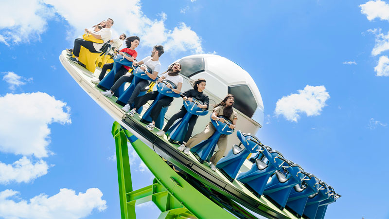 People ride a roller coaster themed with a large soccer ball, set against a bright blue sky with scattered clouds.
